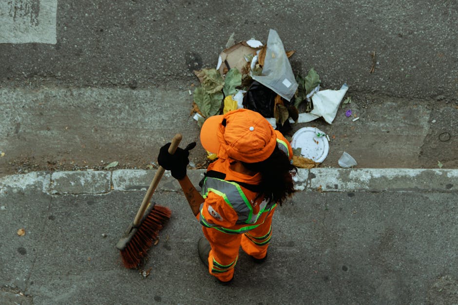 A worker dressed in high-visibility orange clothing and a matching orange cap is standing on a paved pavement next to a concrete wall, engaged in rubbish removal. The worker is holding a long-handled 