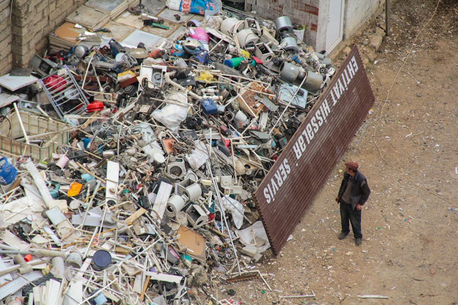 A worker dressed in high-visibility orange clothing and a matching orange cap is standing on a paved pavement next to a concrete wall, engaged in rubbish removal. The worker is holding a long-handled 
